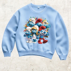 Könnte beinhalten: Hellblaues Sweatshirt mit einem farbenfrohen Aufdruck von mehreren Schlümpfen. Die Schlümpfe sind mit ihrer typischen blauen Haut, weißen Mützen und roten Mützen dargestellt, umgeben von Farbspritzern. Das Sweatshirt hat einen Rundhalsausschnitt und lange Ärmel.