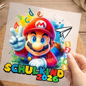 Könnte beinhalten: Ein farbenfrohes Grafikdesign mit Mario, der das Friedenszeichen zeigt. Das Bild enthält den Text "SCHULKIND 2026" in Gelb und Grün, sowie die Zahlen 1-5 und die Buchstaben a-e. Ein Papierflieger ist ebenfalls zu sehen.