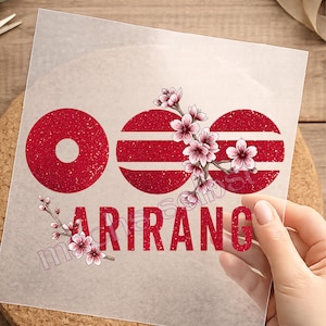Könnte beinhalten: Eine transparente Transferfolie mit einem roten Glitzerdesign, bestehend aus drei kreisförmigen Formen und dem Text ARIRANG in fetter Schrift. Das Design ist mit detaillierten Illustrationen von hellrosa Kirschblütenzweigen und Blüten verziert.