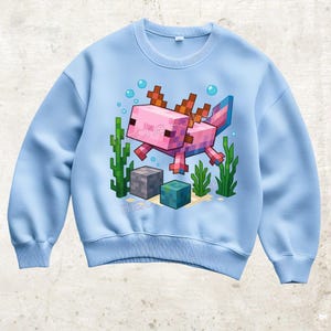 Könnte beinhalten: Hellblaues Sweatshirt mit einem pixeligen rosa Axolotl-Design, braunen und blauen Akzenten, Blasen und Wasserpflanzen. Unten befinden sich zwei pixelige Blöcke. Das Sweatshirt hat einen Rundhalsausschnitt.