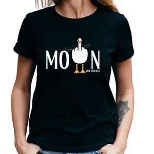 Könnte beinhalten: Schwarzes T-Shirt mit dem weißen Wort "MOON". Ein Cartoon-Gans zeigt den Mittelfinger. Der Text "IHR SPACKEN" steht darunter. Ein humorvolles Design.