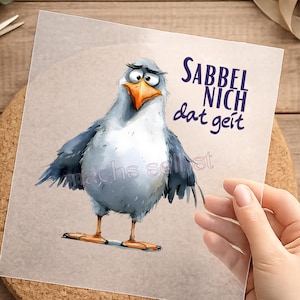 Könnte beinhalten: Ein Druck mit einer Cartoon-Möwe mit traurigem Gesichtsausdruck, grauen und weißen Federn sowie einem orangefarbenen Schnabel und Füßen. Der Text "Sabbel nich dat geit" ist in Dunkelblau geschrieben. Der Druck wird von einer Hand gehalten.