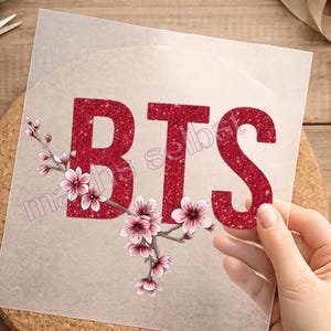 Könnte beinhalten: Ein quadratisches, transparentes Transferblatt mit den Buchstaben BTS in rotem Glitzer-Finish. Ein brauner Zweig mit hellrosa Kirschblüten erstreckt sich über den unteren Teil der Buchstaben. Der Text "maths selbst" ist in Helllila zu sehen.