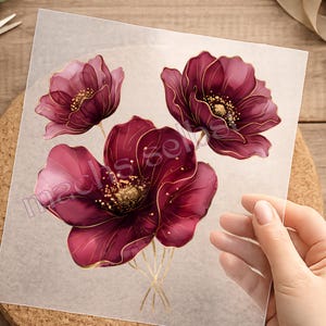 Könnte beinhalten: Ein transparenter Bogen mit einer gedruckten Illustration von drei tiefburgunderroten Blumen mit zarten goldfarbenen Umrandungen und goldenen Staubgefäßdetails. Das Blumenmotiv ist in einer Gruppe mit dünnen, goldenen Stielen angeordnet.