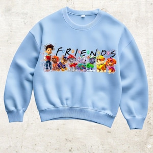 Könnte beinhalten: Hellblaues Sweatshirt mit dem Wort "FRIENDS" und Zeichentrickfiguren. Das Design zeigt einen Jungen und mehrere Hunde in verschiedenen Farben. Das Sweatshirt ist aus einem weichen Material.
