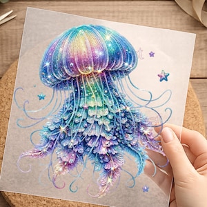 Könnte beinhalten: Ein schillerndes Quallen-Design in Blau-, Lila- und Grüntönen auf transparentem Hintergrund. Das Design zeigt zarte Tentakel und funkelnde Akzente mit Sternformen. Das Bild wird von einer Hand gehalten.