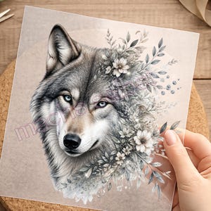 Könnte beinhalten: Ein transparenter Bogen mit einer detaillierten Illustration eines Wolfskopfes mit hellblauen Augen, der teilweise von einem Büschel weißer Blumen und grauer Blätter umgeben ist. Das Design enthält ein dezentes Wasserzeichen über dem Wolfsgesicht.