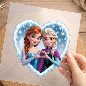 Könnte beinhalten: Ein transparenter Aufkleber mit einem herzförmigen Rahmen aus Eis und Schneeflocken, der eine Illustration von zwei animierten Figuren mit geflochtenem Haar umschließt. Eine Figur hat kastanienbraunes Haar, die andere platinblondes Haar.