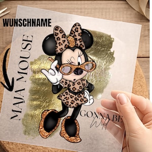 Könnte beinhalten: Eine transparente Transferfolie mit einer Zeichentrickmaus in einem Kleid, einer Schleife und einer Brille mit Leopardenmuster. Das Design enthält einen goldfarbenen Pinselstrich-Hintergrund sowie den Text WUNSCHNAME, MAJA MOUSE und GONNA BE Wild.