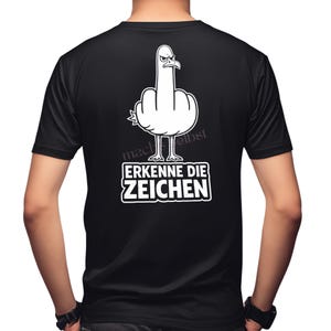 Könnte beinhalten: Ein schwarzes Kurzarm-T-Shirt mit einer weißen Grafik einer Möwe, die eine beleidigende Geste macht. Unter dem Vogel steht der Text "ERKENNE DIE ZEICHEN" in fetten weißen Großbuchstaben. Das Motiv ist mittig auf der Rückseite des Shirts platziert.