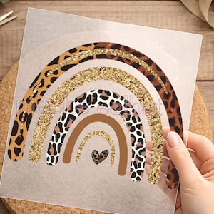 Könnte beinhalten: Ein Regenbogen-Design mit Leopardenmuster, goldenen Glitzerakzenten und einem Herz. Der Regenbogen hat braune, beige und goldene Farben. Das Design befindet sich auf einem transparenten Blatt.