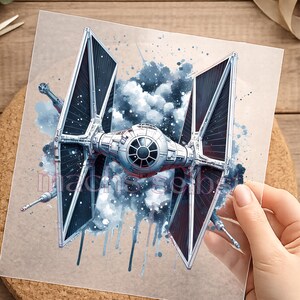 Könnte beinhalten: Detaillierte Illustration eines TIE-Jägers aus Star Wars, mit schwarzen Solarpanelen und einem zentralen Cockpit. Das Kunstwerk enthält blaue Aquarell-Akzente und wird auf einem neutralen Hintergrund präsentiert. Das Bild wird von einer Hand gehalten.