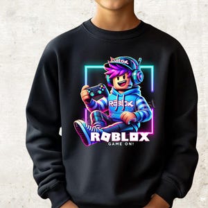 Könnte beinhalten: Ein schwarzes Langarm-Sweatshirt mit einer Grafik einer Cartoon-Figur mit violettem Haar, Kopfhörern und blauem Kapuzenpullover. Die Figur hält einen Game-Controller in einem Neon-Quadrat. Der Text lautet ROBLOX und GAME ON!
