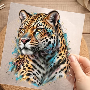 Könnte beinhalten: Eine transparente Transferfolie mit einer detaillierten Illustration eines Leopardenkopfes mit goldgelben Augen, schwarzen Flecken und leuchtend blauen sowie goldenen Farbspritzern. Der Leopard ist im Profil dargestellt und zeigt seine Fellstruktur.