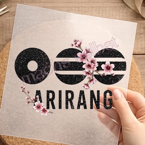 Könnte beinhalten: Eine transparente Transferfolie mit drei schwarzen Glitzerkreisen mit horizontalen Streifen, akzentuiert durch rosa Kirschblütenzweige. Unter den Kreisen ist das Wort ARIRANG in einer fetten, schwarzen Glitzerschriftart gedruckt.