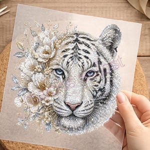 Könnte beinhalten: Ein quadratisches, transparentes Transferblatt mit einer detaillierten Grafik eines weißen Tigerkopfes, der mit silbernen und goldenen Blumenakzenten verziert ist. Der Tiger hat stechend blaue Augen und schwarze Streifen, die Blumen glitzern.