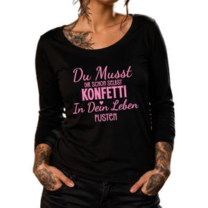 Könnte beinhalten: Ein schwarzes Langarmshirt mit dem in Pink aufgedruckten Text Du Musst dir schon selbst KONFETTI in dein Leben PUSTEN und einem kleinen Herzsymbol. Das Oberteil hat einen Rundhalsausschnitt und besteht aus einfarbigem schwarzem Stoff.