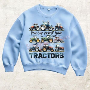 Könnte beinhalten: Hellblaues Sweatshirt mit einer farbenfrohen Grafik verschiedener Traktoren. Der Text "You can never have too many tractors" ist über den Traktoren aufgedruckt. Das Wort "TRACTORS" ist unten in großen Buchstaben aufgedruckt.