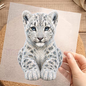 Könnte beinhalten: Ein durchsichtiges Blatt mit einer detaillierten Illustration eines Schneeleoparden-Jungen mit weißem und grauem Fell, dunklen Flecken und leuchtend blauen Augen. Das Junge ist in sitzender Position dargestellt und blickt nach vorne.