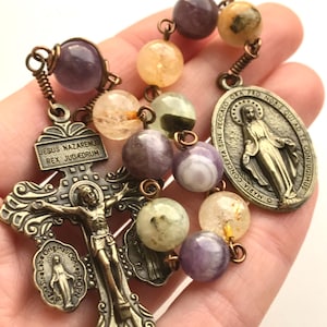 Puede incluir: Un rosario con cuentas de piedras preciosas multicolores y detalles metálicos en tono bronce. El crucifijo presenta la figura de Jesús, con la inscripción "JESUS NAZARENUS REX JUDÆORUM". También hay una medalla de María.