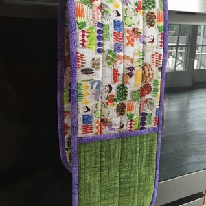May include: A purple and green oven mitt with a colorful print of vegetables and people. The print features a variety of vegetables, including carrots, onions, and peppers. The mitt has a quilted design and a loop for hanging.
