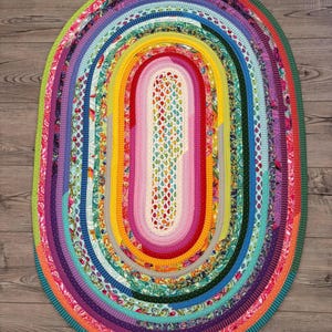 May include: An oval-shaped rug constructed from concentric coils of multi-colored fabric strips. The design features a central section with a small fruit pattern surrounded by alternating bands of solid colors, floral prints, and polka dots.