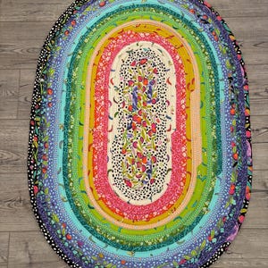 May include: An oval-shaped fabric rug constructed from coiled strips of multi-colored patterned textiles. The design features concentric rings of floral, fruit, and spotted prints in vibrant shades of pink, green, blue, yellow, and black.