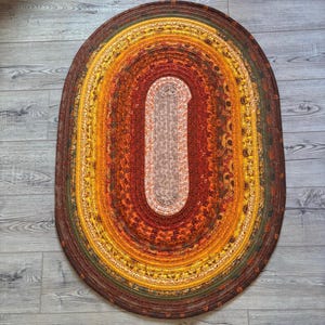 May include: Oval braided rug with a warm color palette. The rug features concentric bands of color, including shades of brown, orange, yellow, and green. The center section is a lighter beige color. The rug is placed on a wooden floor.