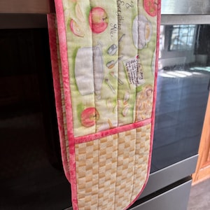 May include: A pink and green oven mitt with a checkered pattern on the bottom. The mitt has a pocket on the back and features a print of apples, a pot, and a recipe card that says "Spice & Everything Nice".