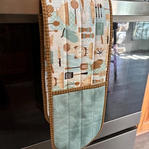 May include: A brown and blue quilted oven mitt with a pattern of kitchen utensils and a pocket on the bottom. The mitt is hanging on a silver oven door.