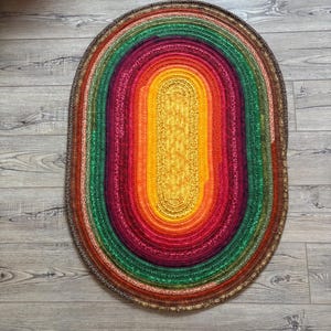 May include: Oval-shaped, multi-colored rug with a rainbow design. The rug features concentric bands of color, including red, orange, yellow, green, and purple. The rug is made of fabric strips and has a brown border. The rug is placed on a wooden floor.