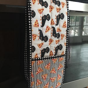 May include: A black and white oven mitt with a pattern of black dachshunds and pizza slices. The mitt has a black and white polka dot trim and a black and white striped pocket.