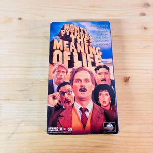 May include: A VHS video cassette of the film "Monty Python's The Meaning of Life." The cover shows a collage of actors with surprised expressions, the film's title, and the MCA Universal logo.
