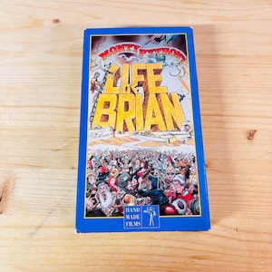May include: A vintage VHS tape of the film "Monty Python's Life of Brian." The cover features colorful illustrations of a crowd of people and the film's title in large, yellow letters. The tape is in a blue case.