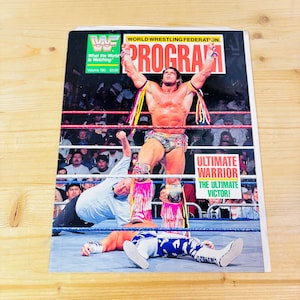 Puede incluir: Una portada de revista vintage del programa de la Federación Mundial de Lucha Libre. La portada presenta a un luchador con los brazos levantados y a otro luchador en la lona. La revista tiene el texto "PROGRAM" y "ULTIMATE WARRIOR THE ULTIMATE VICTOR!"
