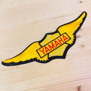 May include: Yellow and black Yamaha patch with a wing design. The patch features the word "YAMAHA" in red letters on a black rectangle. The wings are yellow with black outlines.