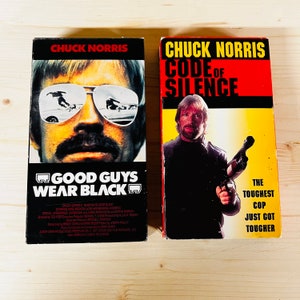 May include: Two VHS tapes featuring Chuck Norris. One tape is titled "Good Guys Wear Black" with Norris wearing sunglasses. The other tape is titled "Code of Silence" with Norris holding a gun. Both tapes have black, red, and yellow color schemes.