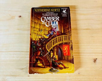 May include: A paperback book cover with a yellow background and a drawing of a group of people in medieval clothing on a staircase. The title of the book is "Camber of Culdi" and the author is "Katherine Kurtz".