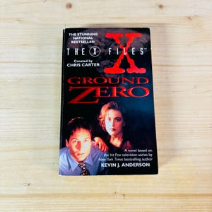 May include: A paperback book cover with a black background and red text. The title is "The X Files" and "Ground Zero". The cover features a photo of two people, a man and a woman, with the woman in the foreground. The text on the cover says "A novel based on the hit Fox television series by New York Times bestselling author Kevin J. Anderson."