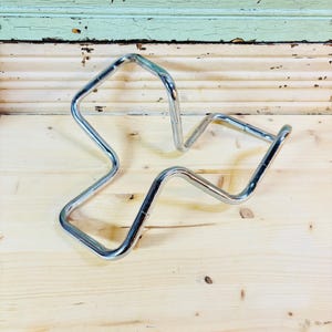 May include: A chrome-plated metal wine bottle holder with a unique, abstract design. The holder is shaped with curved lines and angles, creating an open, geometric form. It sits on a light-colored wooden surface.