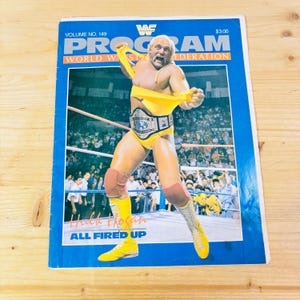 Puede incluir: Una revista vintage del programa de la WWF con Hulk Hogan con un atuendo de lucha libre amarillo y rojo. La portada tiene el texto "PROGRAM" y "WORLD WRESTLING FEDERATION". La revista es azul con un borde blanco y el texto "ALL FIRED UP".