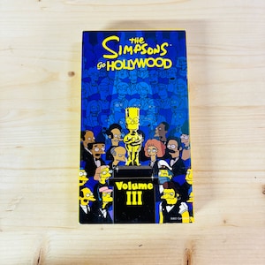 May include: A DVD box set for "The Simpsons Go Hollywood Volume III" featuring Bart Simpson holding an Oscar statue. The cover is predominantly blue with a crowd of cartoon characters in the background.