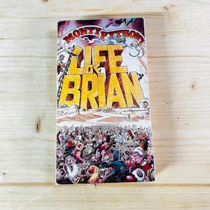 Może przedstawiać: Kasetę VHS z filmem "Żywot Briana" Monty Pythona na drewnianej powierzchni. Okładka przedstawia tytuł dużymi żółtymi literami, ze sceną tłumu i ilustracjami. Kaseta ma kształt prostokąta.