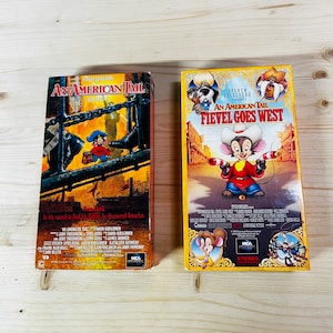 Puede incluir: Dos cintas VHS de la película de animación "An American Tail" y su secuela "Fievel Goes West". La primera cinta tiene un fondo oscuro con un personaje de ratón. La segunda cinta tiene un fondo amarillo con un personaje de ratón con sombrero de vaquero.