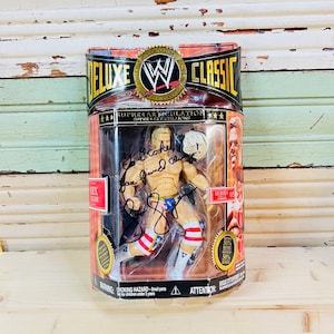 Może przedstawiać: Figurka akcji WWE Deluxe Classic Lexa Lugera w przezroczystym plastikowym opakowaniu. Figurka ma na sobie czerwony, biały i niebieski strój do zapasów i pas mistrzowski. Opakowanie zawiera logo WWE i napis "Series 03".