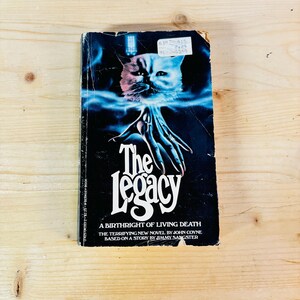 May include: A paperback book cover with a black background and a white cat with blue eyes. The title of the book is "The Legacy: A Birthright of Living Death" by John Coyne, based on a story by Jimmy Sangster.
