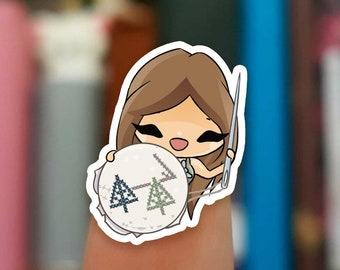 Puede incluir: Una pegatina de una niña de dibujos animados que sostiene una aguja e hilo, trabajando en un proyecto de punto de cruz con un diseño de dos árboles verdes.
