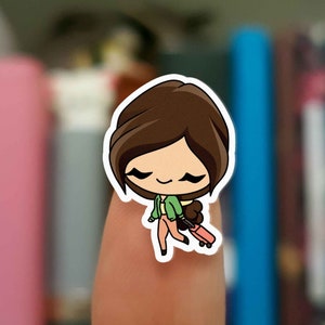May include: A sticker featuring a cartoon character with brown hair, wearing a green jacket and peach pants, carrying a pink suitcase. The character is smiling with closed eyes, suggesting a travel theme.