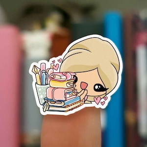 Puede incluir: Una pegatina de dibujos animados de una mujer rubia con una pila de libros y bolígrafos en una taza. La mujer sonríe y tiene corazones a su alrededor.