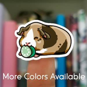 May include: A sticker featuring a cartoon guinea pig with brown, white, and tan fur, holding a green cucumber slice. The text "More Colors Available" is displayed in white at the bottom of the image.